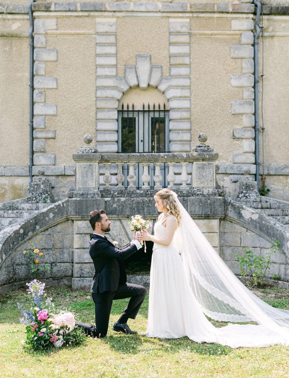 Marié agenouillé offrant le bouquet à la mariée devant l’escalier en fer à cheval du château de Motteux
